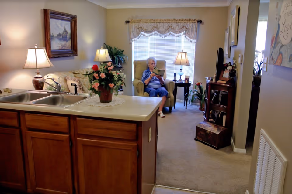 A cozy living room area with a woman sitting in an armchair reading a book. The room has beige walls, carpeted floor, and is softly lit by table lamps. A kitchen counter with a double sink and a flower pot is visible in the foreground. There are framed pictures and plants decorating the space.