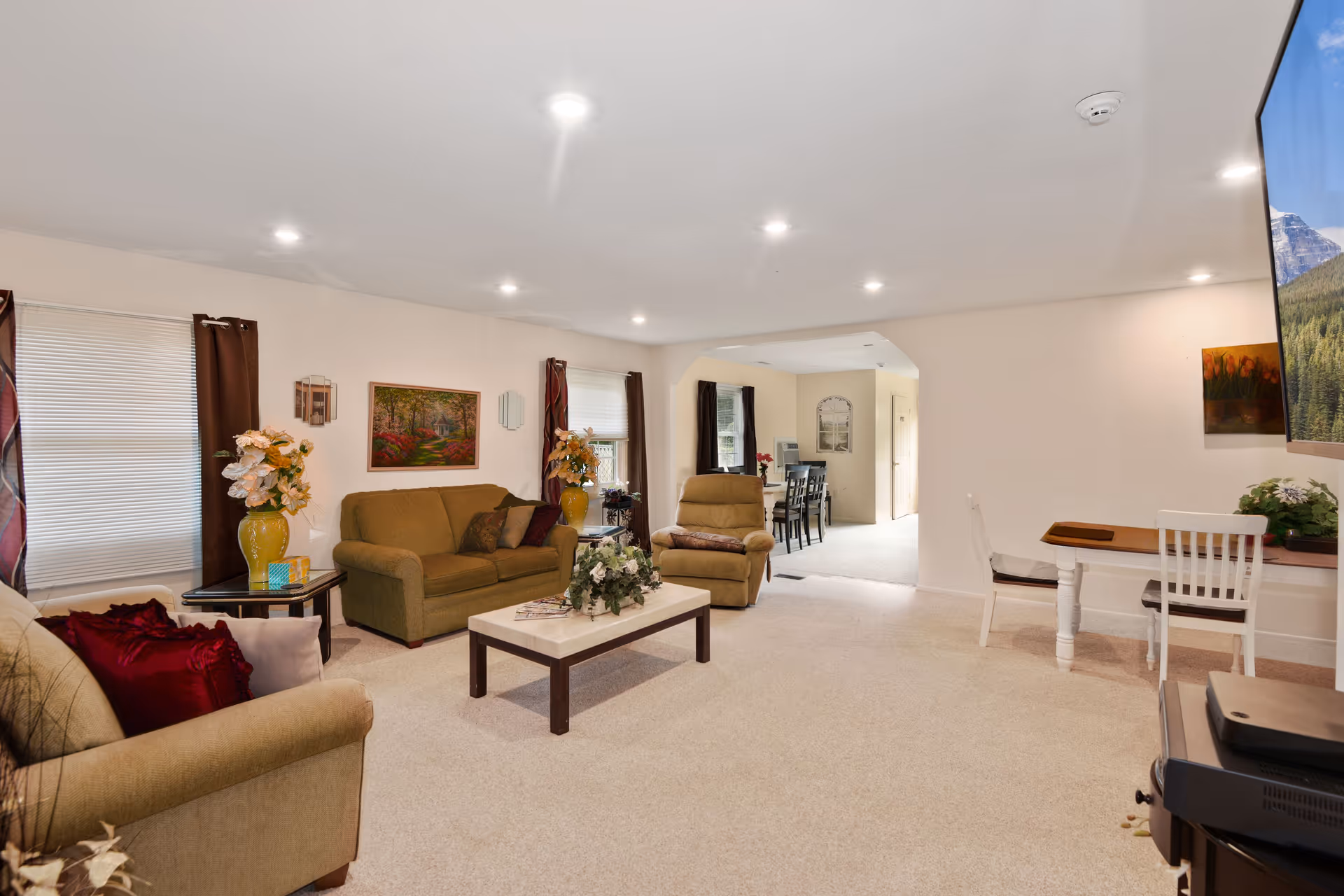 A spacious living room with beige carpet, two green sofas, a beige armchair, a white coffee table with a floral centerpiece, and a small desk with two white chairs. The room is decorated with paintings, vases with flowers, and has multiple windows with brown curtains. In the background, a dining area with a dark wooden table and chairs is visible.