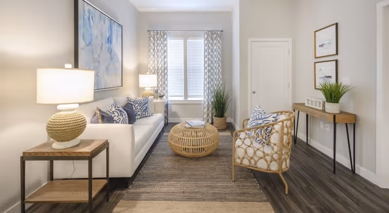 Bright modern living room with a white sofa, rattan chair and round wicker coffee table, a console table, lamps and a window with patterned curtains.