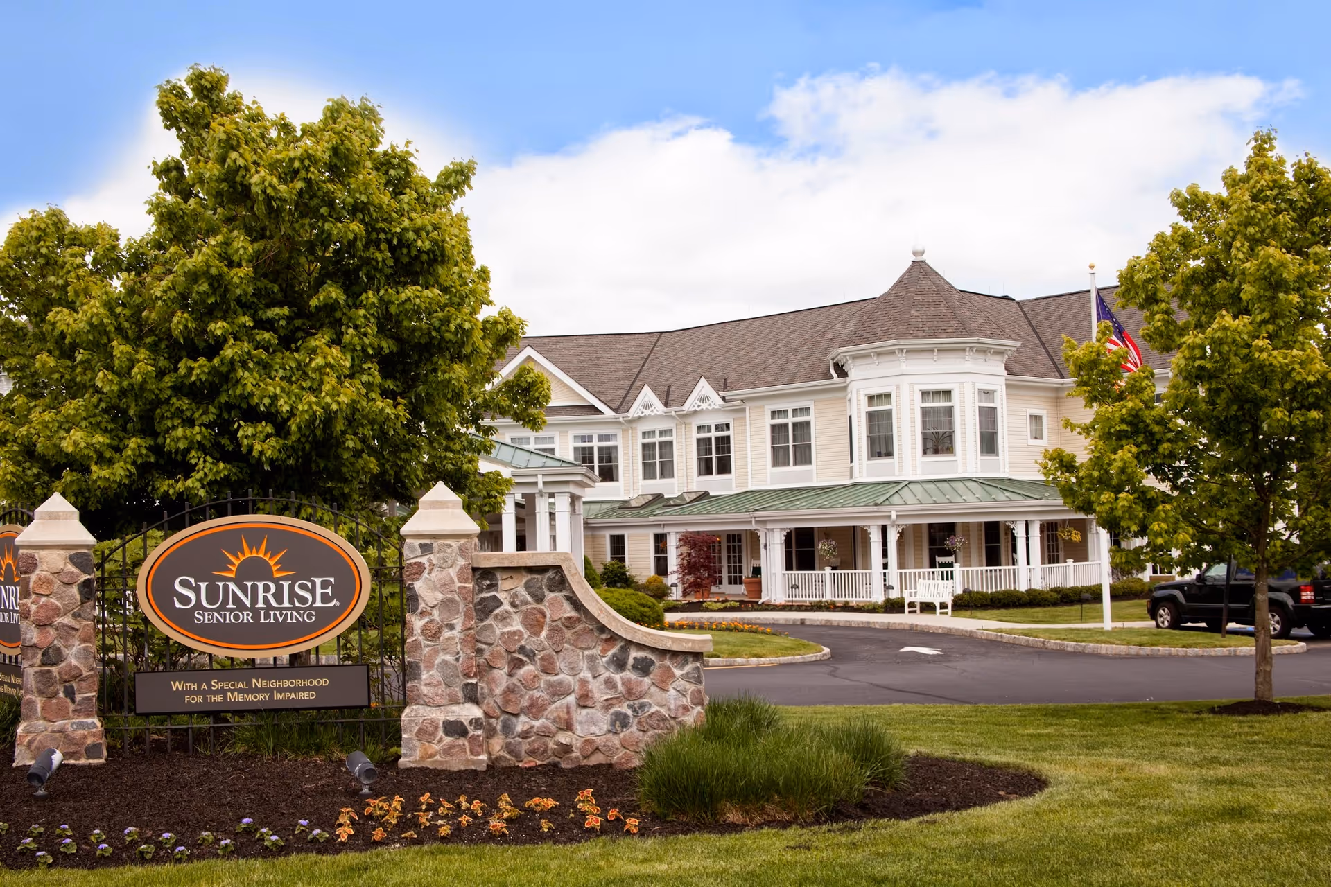 Exterior view of Sunrise Senior Living facility showing a large, two-story building with white siding and a green roof. The building has multiple windows and a covered porch with white railings. In front, there is a stone sign with the Sunrise Senior Living logo and the text 'With a Special Neighborhood for the Memory Impaired.' The area is landscaped with grass, trees, and flowers under a partly cloudy sky.