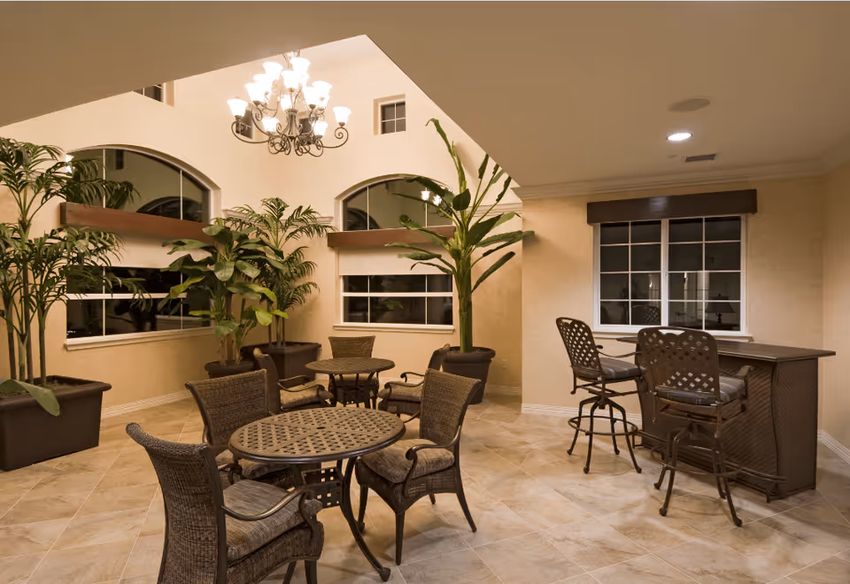 Indoor seating area with round tables and wicker chairs, large potted plants, a chandelier hanging from a high ceiling, and a bar counter with two high chairs near a window.