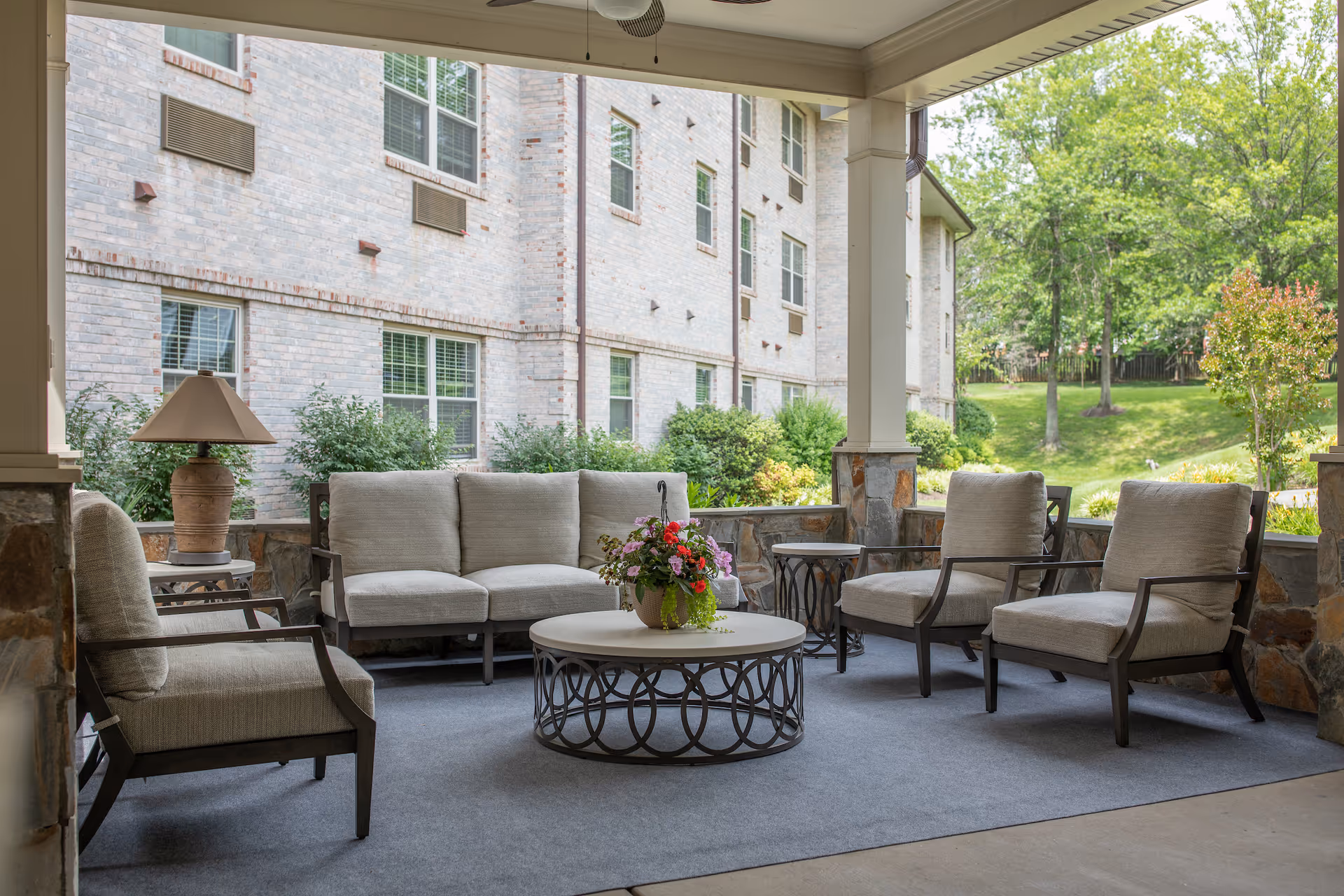 Covered outdoor seating area with cushioned chairs and a sofa arranged around a round coffee table with a flower pot on top. The area is carpeted and overlooks a green lawn with trees and shrubs. The building's brick exterior is visible in the background.