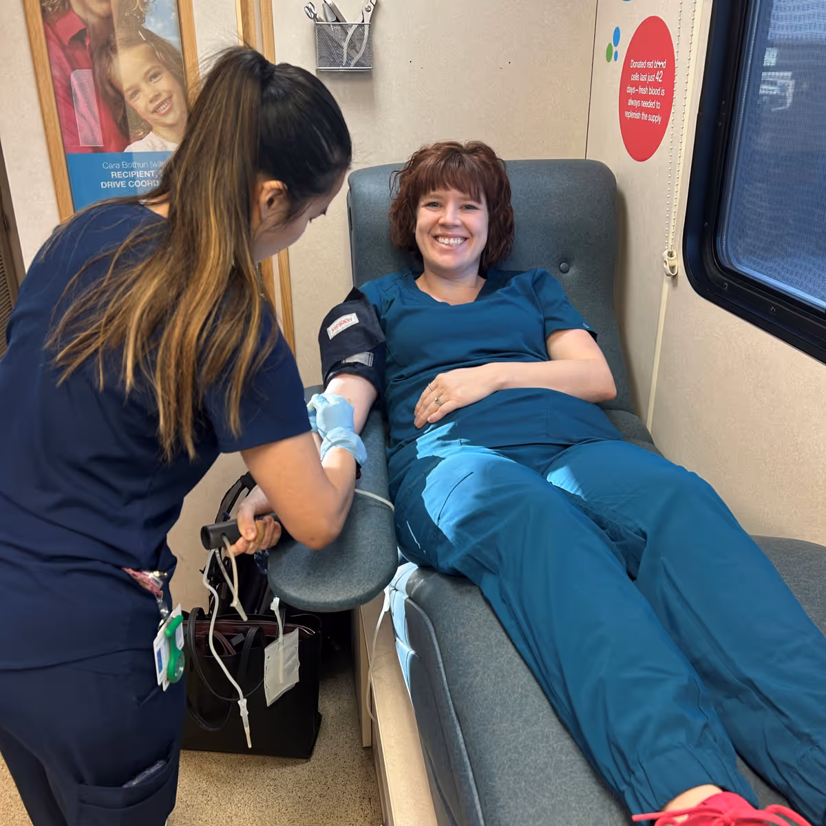 A woman in teal scrubs is lying on a medical recliner chair smiling while a healthcare worker in navy scrubs is preparing to draw blood from her arm inside a medical room.