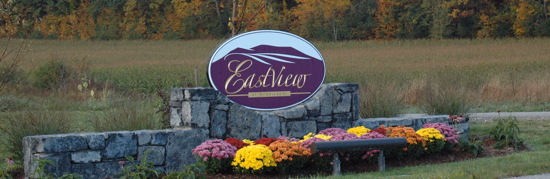 Stone entrance sign reading "EastView at Middlebury" surrounded by a low stone wall and a bed of colorful flowers.