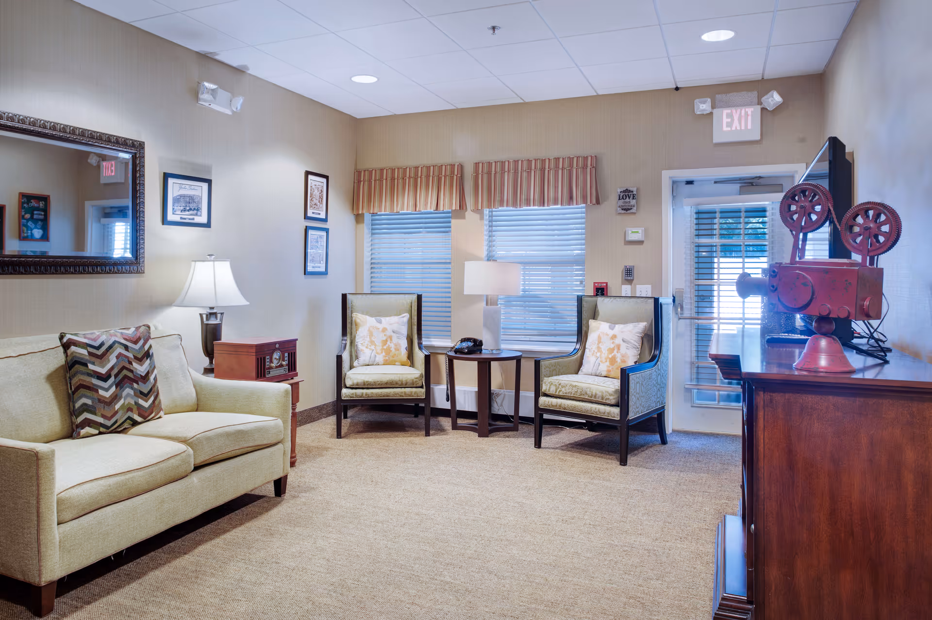 A cozy living room area with a beige sofa adorned with patterned pillows, two cushioned armchairs with floral pillows, a wooden side table with a lamp and a telephone, and a wooden cabinet holding a vintage red film projector. The room has beige walls, a large mirror, framed pictures, two windows with blinds and valances, and an exit door with a glass panel. The ceiling has recessed lighting.