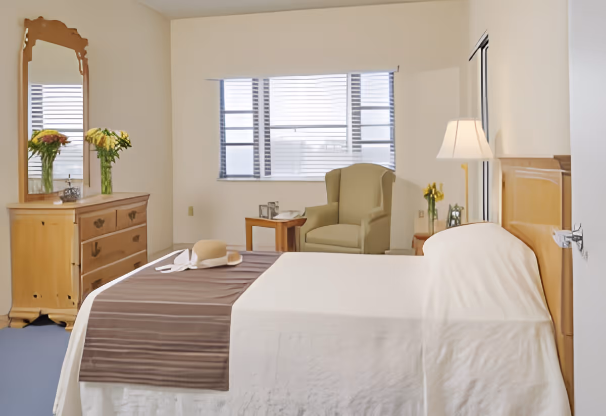 A bright bedroom with a neatly made bed covered with a white bedspread and a brown runner. A beige armchair sits near a window with blinds, next to a small wooden side table holding a glass and a book. A wooden dresser with a large mirror and two vases of flowers is on the left side of the room. A floor lamp is positioned next to the bed on the right side.