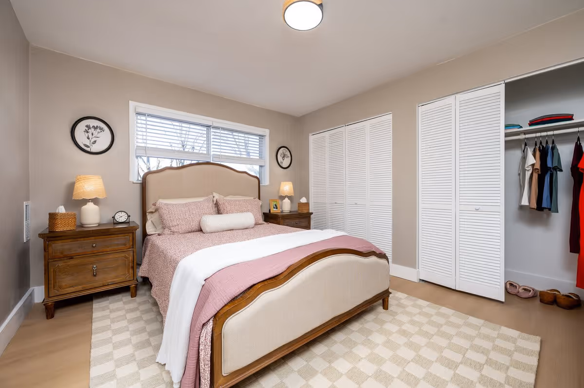 A neatly arranged bedroom with a wooden bed frame featuring beige upholstery and pink bedding. There are two wooden nightstands on either side of the bed, each with a lamp and decorative items. A window with white blinds is above the bed, and a closet with white louvered doors is visible on the right side, partially open showing hanging clothes and shoes. The room has beige walls and a checkered beige and white rug on the floor.