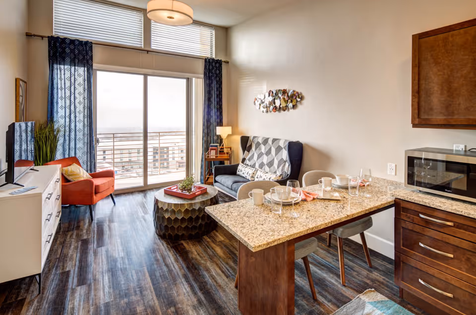 A modern living area with a small kitchen counter set for dining, a dark blue couch with a geometric patterned throw, an orange armchair, a round coffee table, a TV on a white stand, and large sliding glass doors with blue curtains leading to a balcony with a city view.
