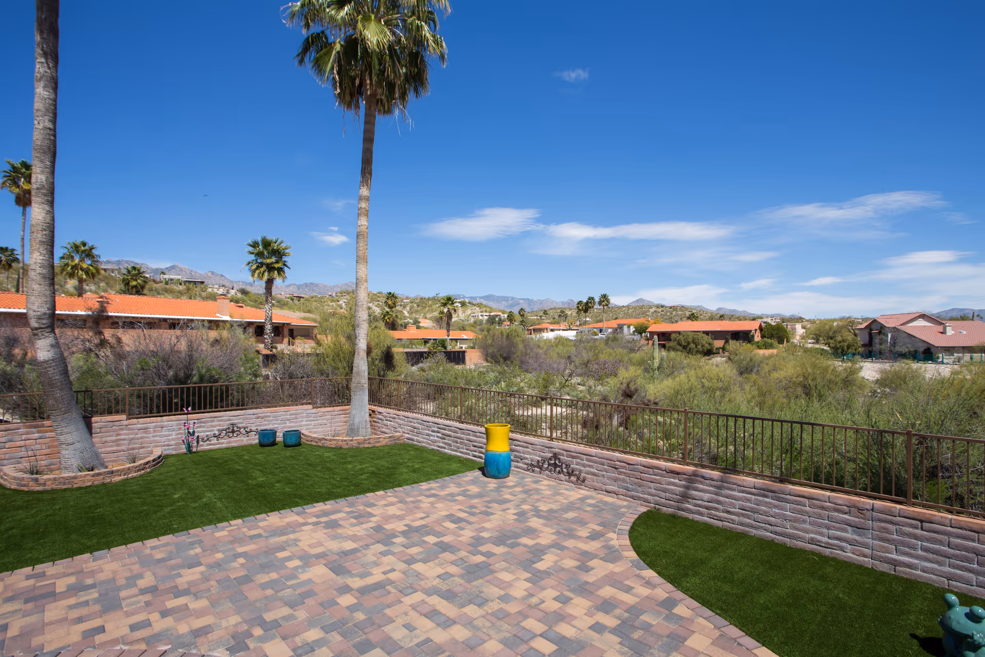 Outdoor patio area with a paved brick floor and artificial grass patches, surrounded by a low brick wall and metal railing. Several tall palm trees are visible, along with desert vegetation and houses with red-tiled roofs in the background under a clear blue sky.