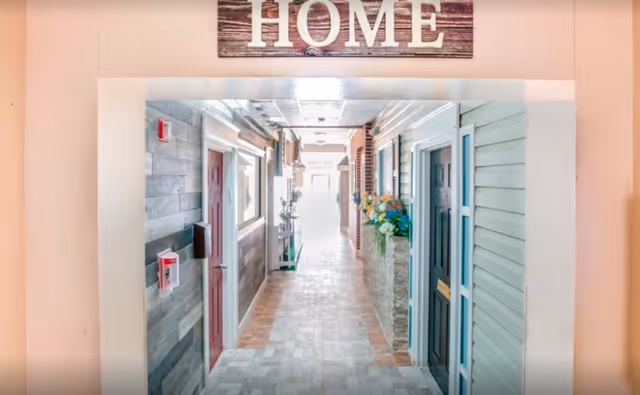 Indoor hallway of a senior living facility with multiple doors on both sides, decorated with flowers and a wooden sign above the entrance that reads 'HOME'.