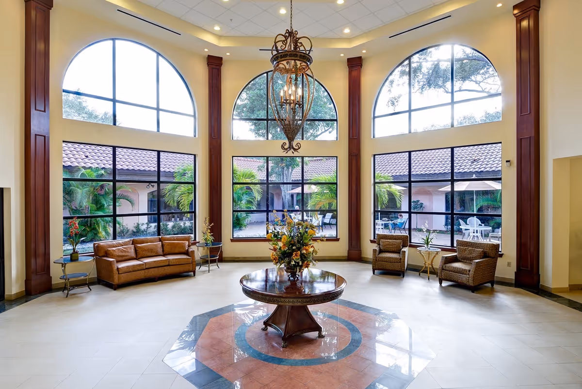 Spacious and elegant common area with large arched windows letting in natural light, a central round wooden table with a floral arrangement, a brown leather sofa on the left, two wicker armchairs with a small table on the right, and a decorative chandelier hanging from the ceiling.