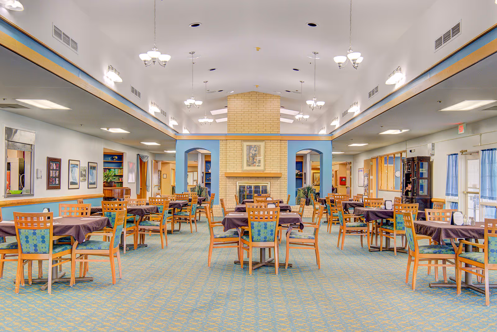 A spacious dining room in an assisted living facility with multiple wooden tables and chairs arranged neatly. The tables are covered with dark tablecloths, and the chairs have patterned cushions. The room features a central brick fireplace, light blue walls with wooden trim, and several chandeliers hanging from the ceiling. Large windows with blue curtains allow natural light to enter the room.