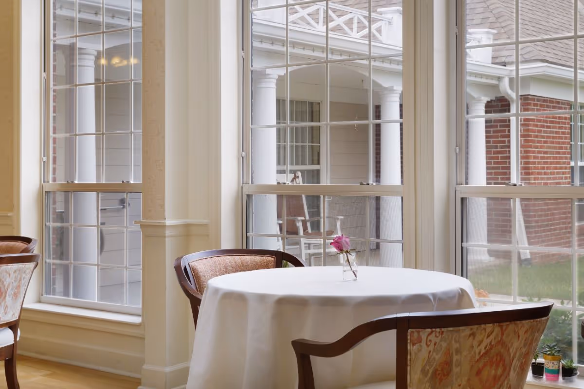 A round table covered with a white tablecloth is set near large windows with a view of a porch with white columns and brick walls. The table has a small vase with a single pink rose. Two upholstered chairs with wooden frames are positioned around the table.