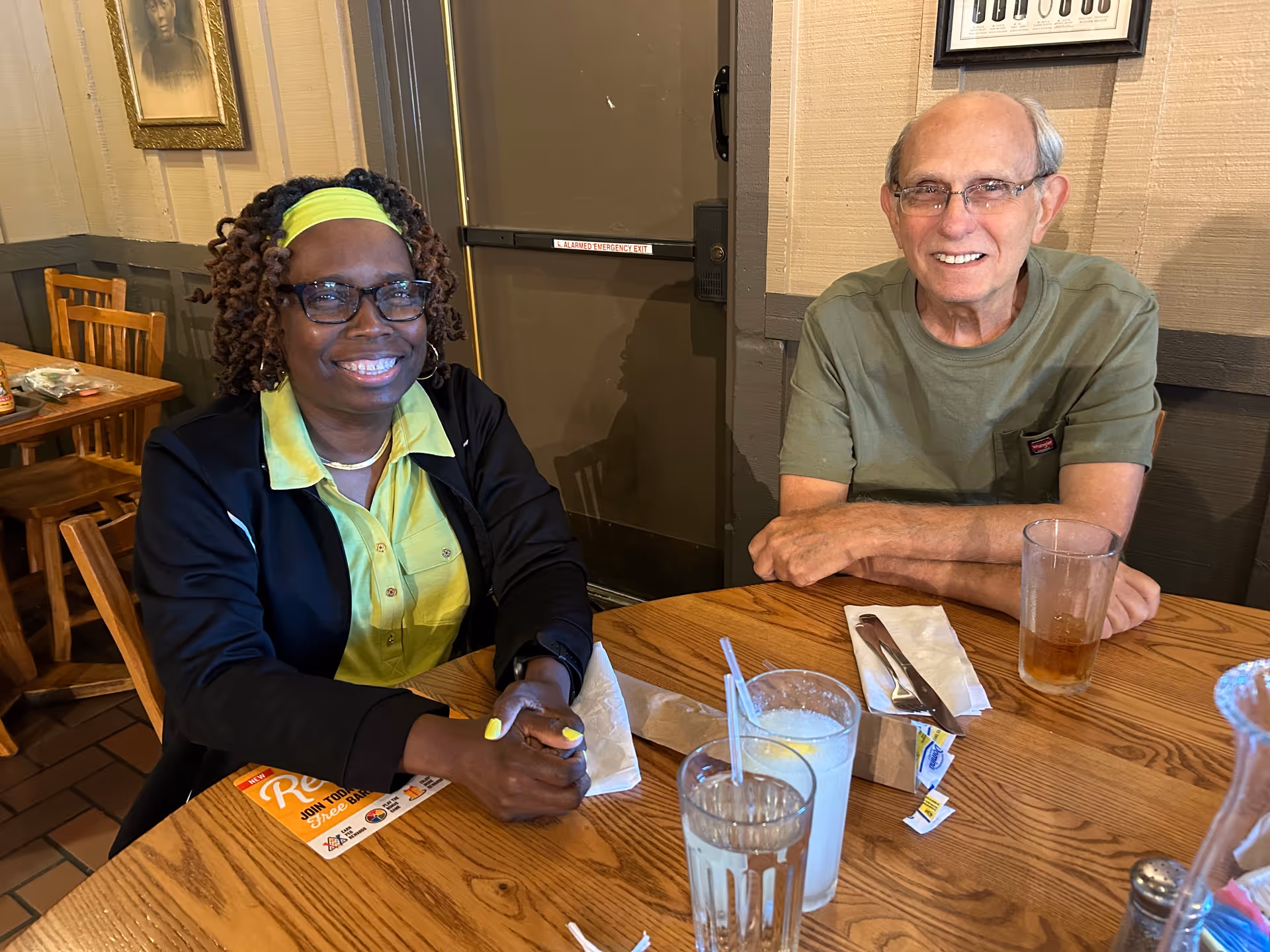 Two people sitting at a wooden table in a dining area. The person on the left is a woman with curly hair wearing glasses, a yellow headband, and a black jacket over a yellow shirt. The person on the right is an older man with glasses wearing a green t-shirt. On the table are two glasses of water, a glass with iced tea, napkins, utensils, and a small packet. The background shows wooden chairs, a door, and framed pictures on the wall.