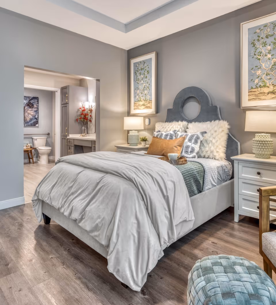 A cozy bedroom with a neatly made bed featuring a gray headboard, white and patterned pillows, and a light gray comforter. Two bedside tables with lamps flank the bed. The room has wooden flooring and gray walls adorned with framed botanical artwork. An open doorway reveals a bathroom with a toilet and a vanity with a vase of red flowers.