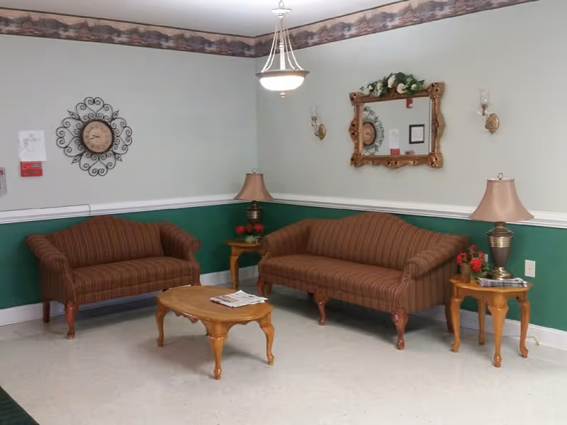 A sitting area with two brown striped sofas facing a wooden coffee table with newspapers on it. There are two wooden side tables with lamps and small flower arrangements. A decorative wall clock and a framed mirror with floral decoration are mounted on the light green walls with a darker green lower half and a wallpaper border near the ceiling.