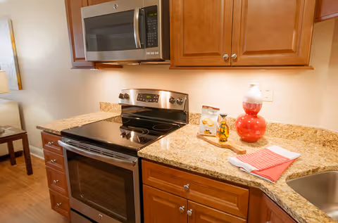 A kitchen area with wooden cabinets, a stainless steel electric stove and oven, a microwave above the stove, granite countertops, a red decorative vase, a small bottle of olive oil, a cutting board with a snack bag, and a folded red and white kitchen towel next to a sink.