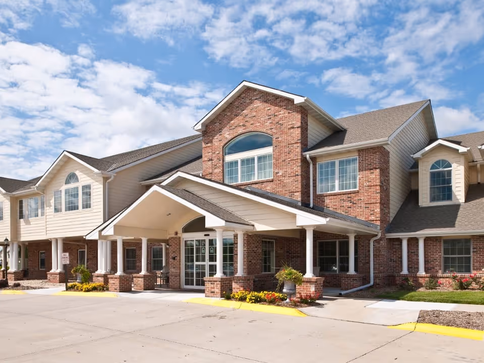 Exterior view of a senior living facility building with a brick and beige siding facade, large windows, and a covered entrance with white columns. The sky is partly cloudy and the driveway is visible in front.