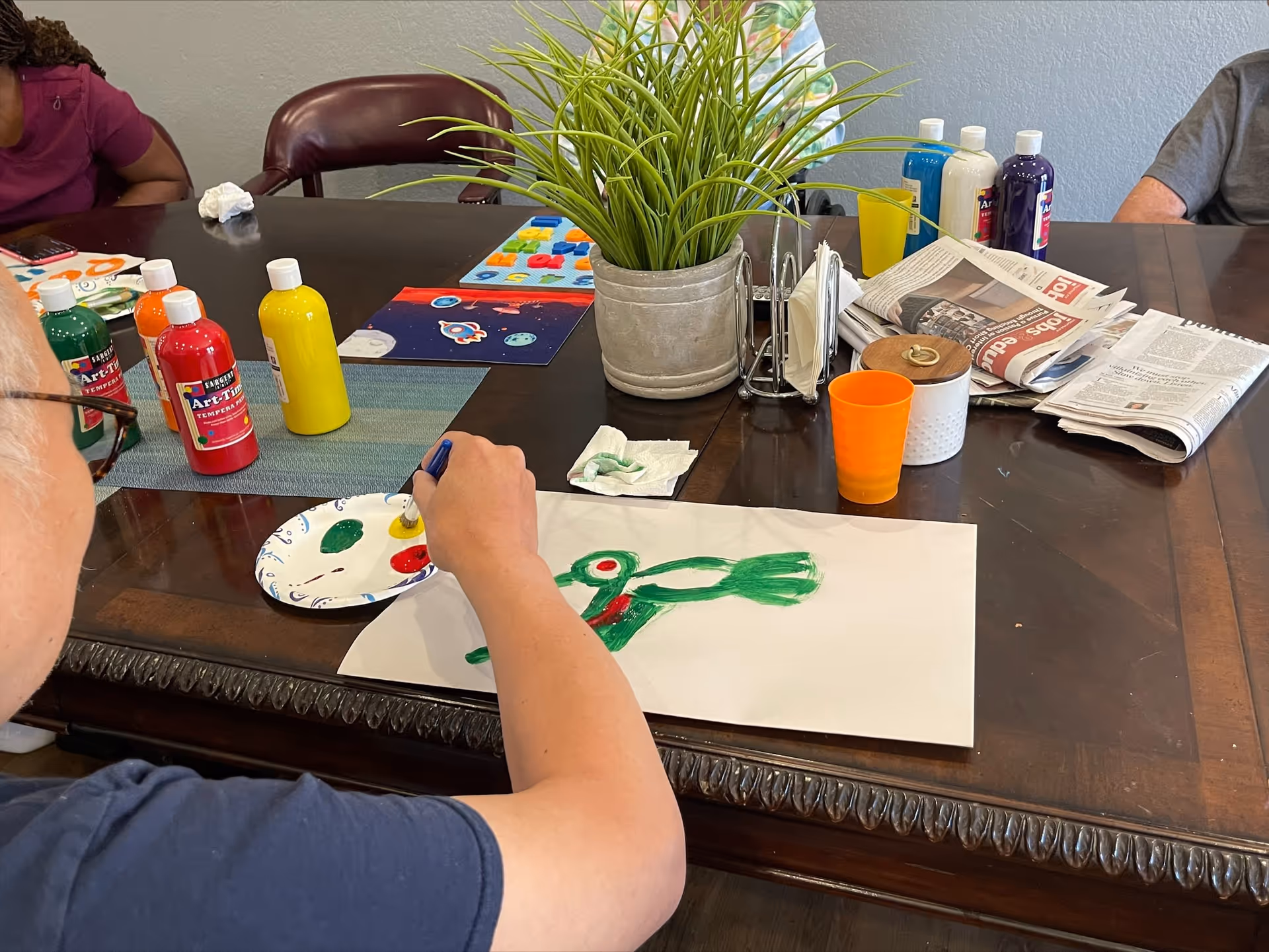 A person painting a green bird on a white sheet of paper at a wooden table. Various bottles of paint in red, yellow, green, blue, and purple are on the table along with a paper plate used as a palette, a potted plant, newspapers, and an orange plastic cup. Other people are partially visible around the table.