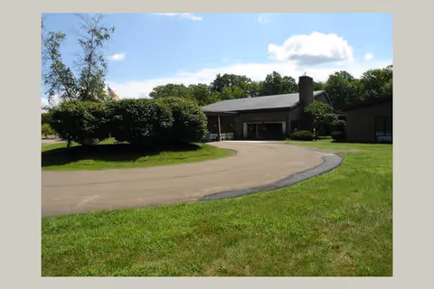 A single-story building with a covered entrance and a curved driveway surrounded by green grass and bushes under a partly cloudy sky.