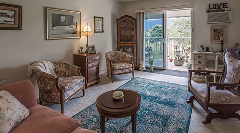 A cozy living room with floral upholstered chairs, a pink sofa with floral pillows, a wooden coffee table on a blue patterned rug, and a small wooden cabinet with a lamp. There is a sliding glass door leading to a balcony with plants and a view of greenery and mountains. The room has framed pictures on the walls and a decorative 'LOVE' sign above an air conditioner unit.