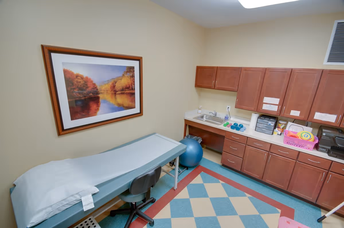 A small medical or therapy room with a padded examination table covered with a disposable sheet, a rolling stool, and a large framed landscape picture on the wall. There are wooden cabinets and a countertop with a sink, small dumbbells, a blue exercise ball, and various office and therapy supplies. The floor has a colorful checkered pattern.