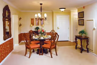 A dining area in a senior living community featuring a round wooden table set for four with floral centerpiece and orange cushioned chairs. The room has beige walls and carpet, a chandelier overhead, a decorative mirror on the left wall, framed artwork, and a small wooden side table with a plant on the right.