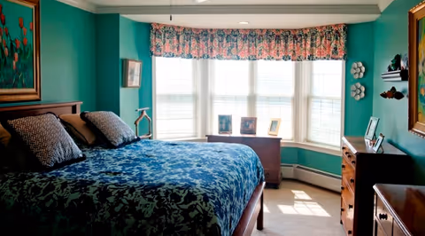 A bedroom with teal walls and a large window with floral valance letting in natural light. The room features a bed with a blue and black patterned bedspread and two pillows, wooden dressers with framed photos and decorative items, and framed artwork on the walls.