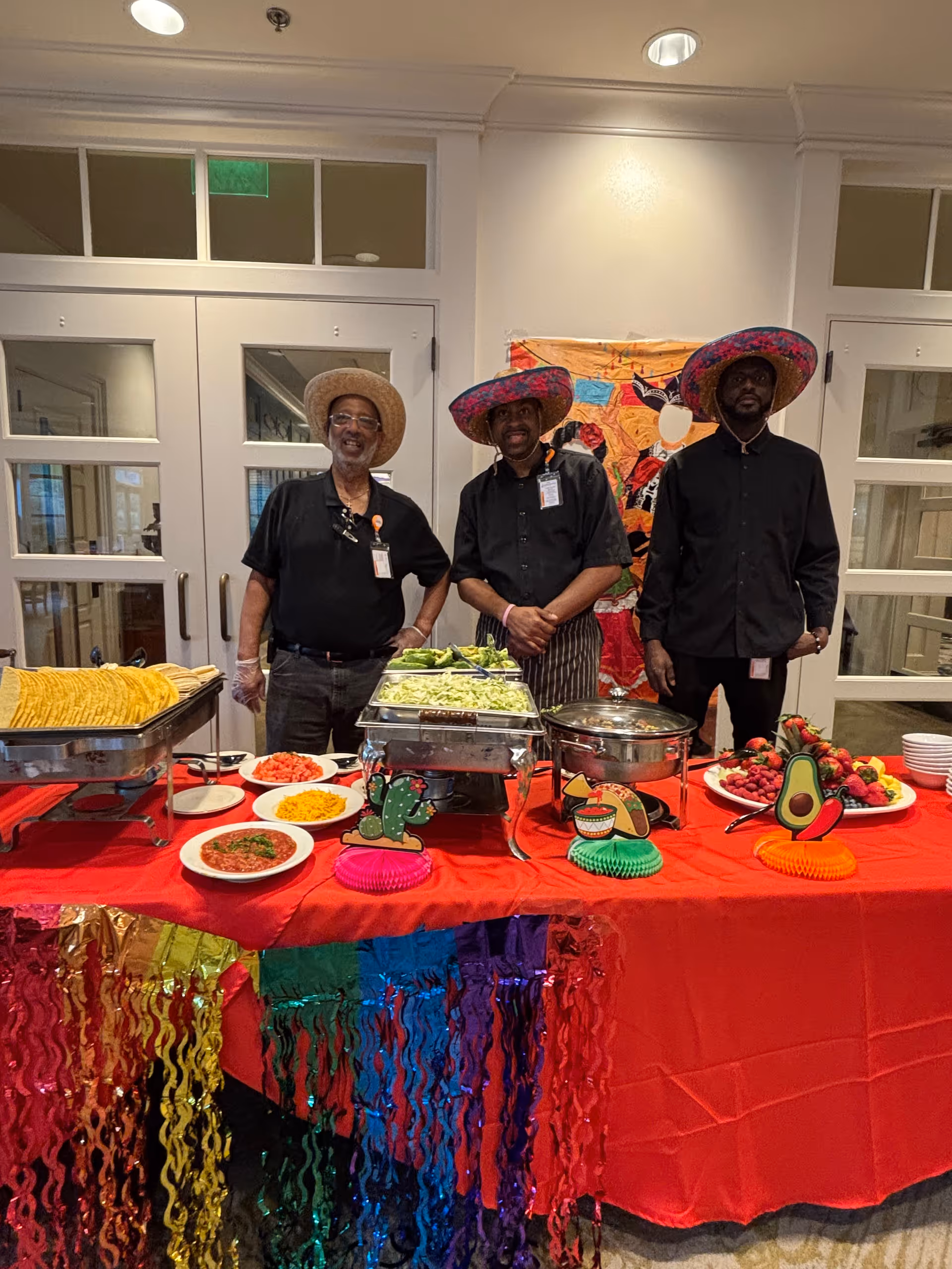 Three men wearing black shirts and colorful sombreros stand behind a buffet table decorated with a red tablecloth and colorful hanging streamers. The table has various food items including tortillas, chopped vegetables, and sauces, with festive decorations like a cactus, taco, and avocado cutouts. The setting appears to be indoors with double glass doors in the background.