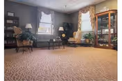 A cozy living room with beige patterned carpet, two windows with curtains, a green sofa, a yellow armchair, a wooden coffee table, a glass display cabinet, and various plants and lamps providing a warm atmosphere.