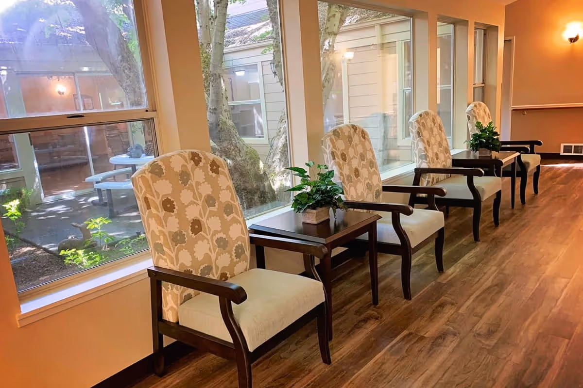 Row of upholstered armchairs and small side tables lined along large windows overlooking a shaded courtyard.
