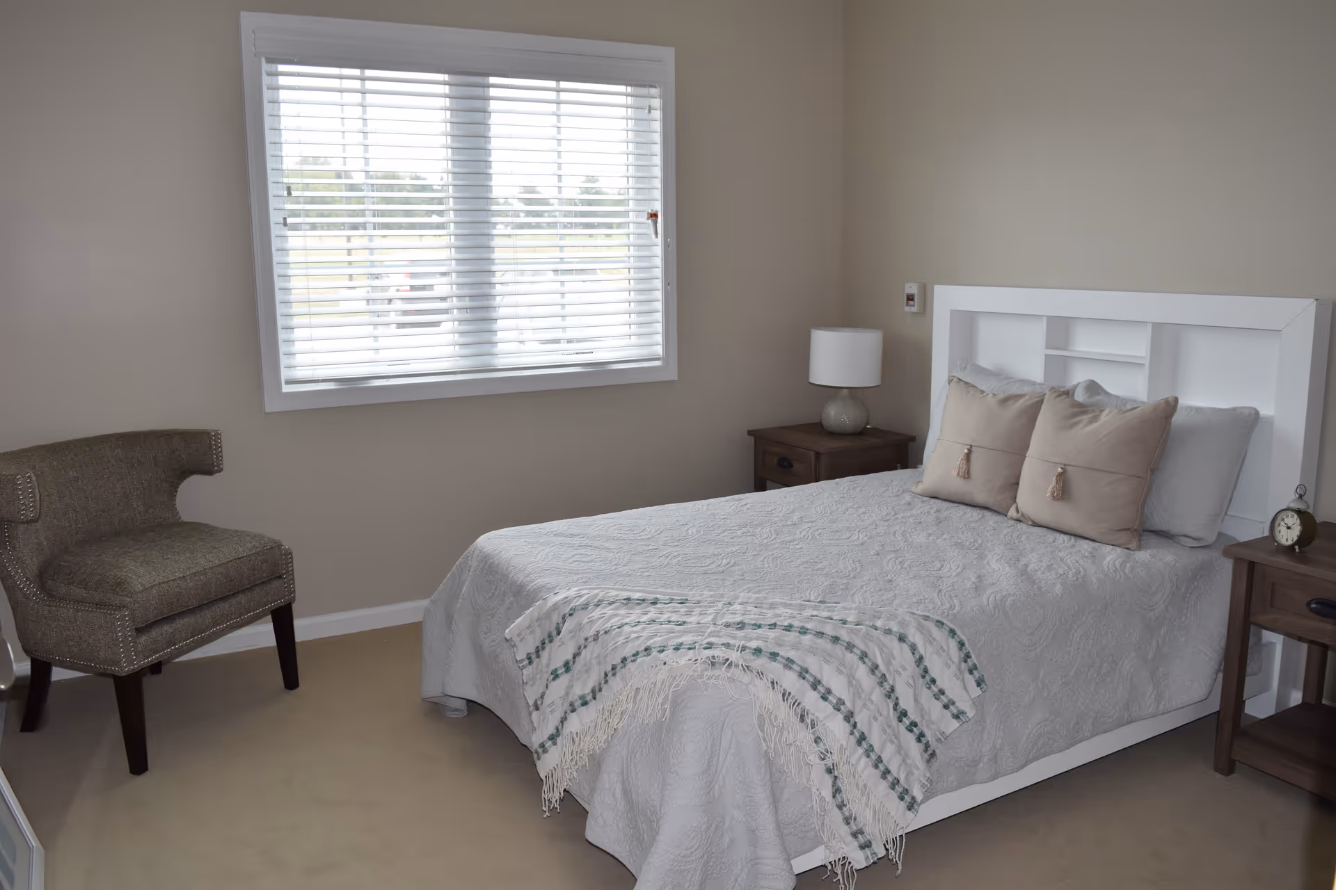 A bedroom with a white bed frame, a bed covered with a light gray quilt and two beige pillows. A striped throw blanket is draped over the foot of the bed. There are two wooden nightstands on either side of the bed, one with a lamp and the other with a small clock. A gray upholstered chair is positioned near a window with white blinds, and the walls are painted beige.