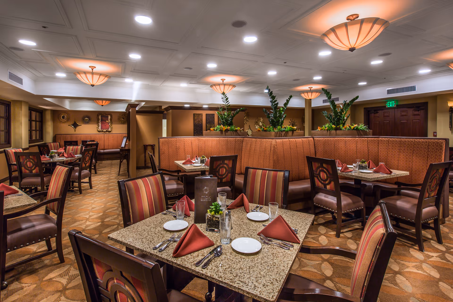 Interior view of a dining room with multiple tables set with plates, glasses, silverware, and folded red napkins. The room features upholstered chairs with striped fabric, booth seating with patterned cushions, decorative plants on dividers, warm lighting fixtures on the ceiling, and wall decorations.