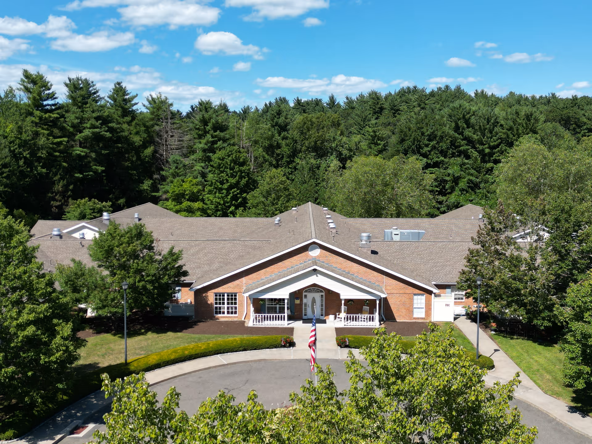 Front exterior view of Arden Courts - ProMedica Memory Care Community (Avon), a single-story brick building with a covered entrance, surrounded by trees and greenery under a blue sky with scattered clouds.
