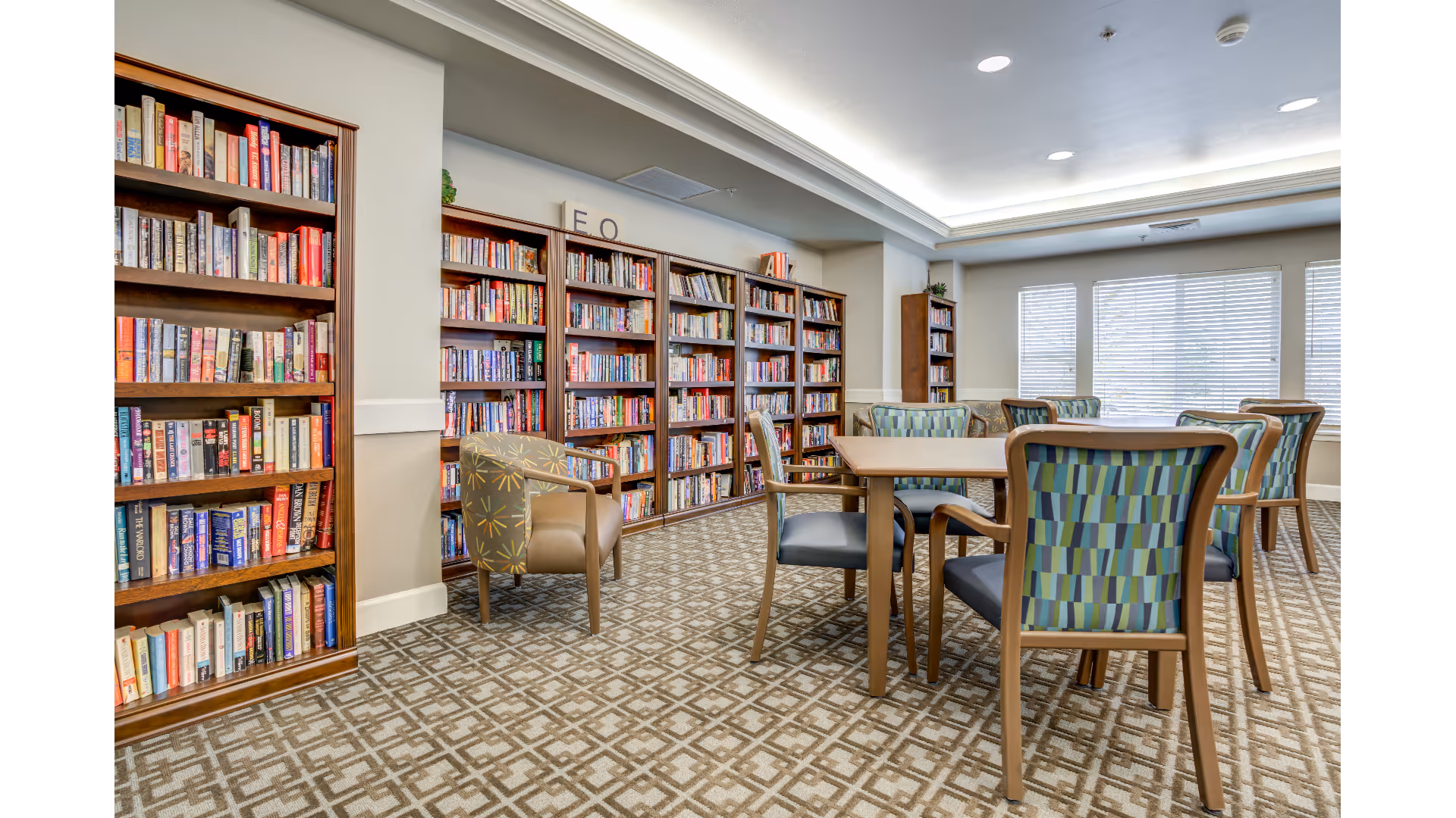 A well-lit room with multiple wooden bookshelves filled with books along the walls. There are several tables with patterned chairs arranged around them, and a single armchair with a different pattern near the bookshelves. Large windows with blinds allow natural light to enter the room. The carpet has a geometric pattern, and the ceiling has recessed lighting.