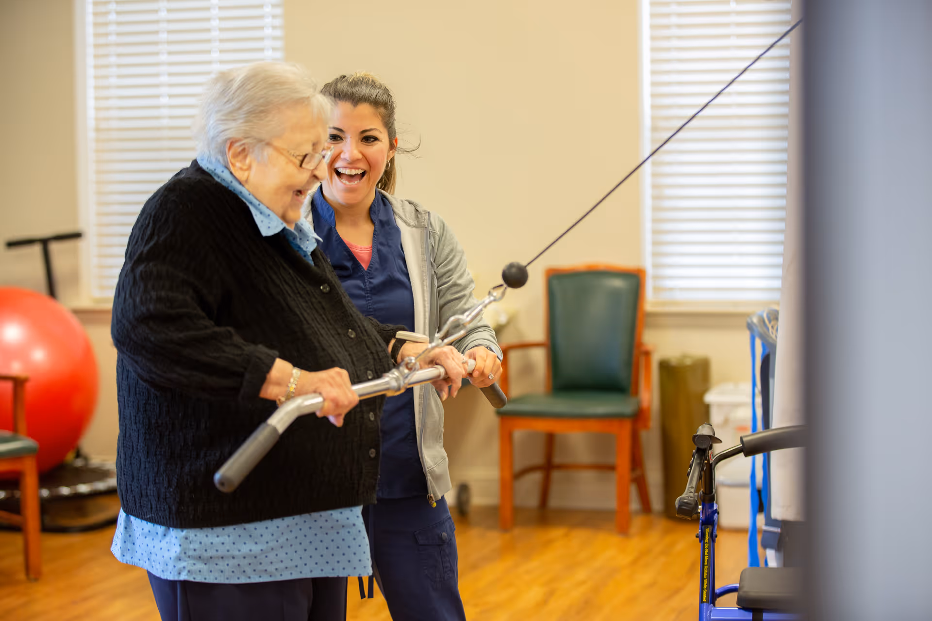 An elderly woman is exercising with a cable machine while a caregiver or physical therapist stands beside her, smiling and providing support in a room with wooden floors, chairs, and a large red exercise ball in the background.