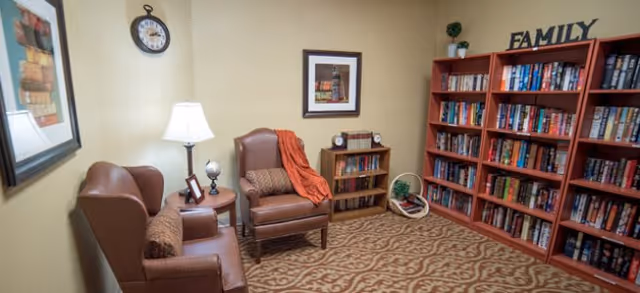 A cozy reading room with two brown leather armchairs, a small round wooden table with a lamp and picture frames, a bookshelf filled with books, and a larger bookshelf labeled 'FAMILY' also filled with books. The room has beige walls, a patterned carpet, and framed artwork on the walls.