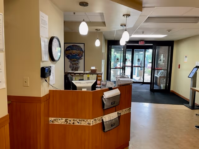Reception desk area inside Buckeye Forest at Fairfield facility with a clock on the wall, hanging pendant lights, and glass double doors leading outside in the background.