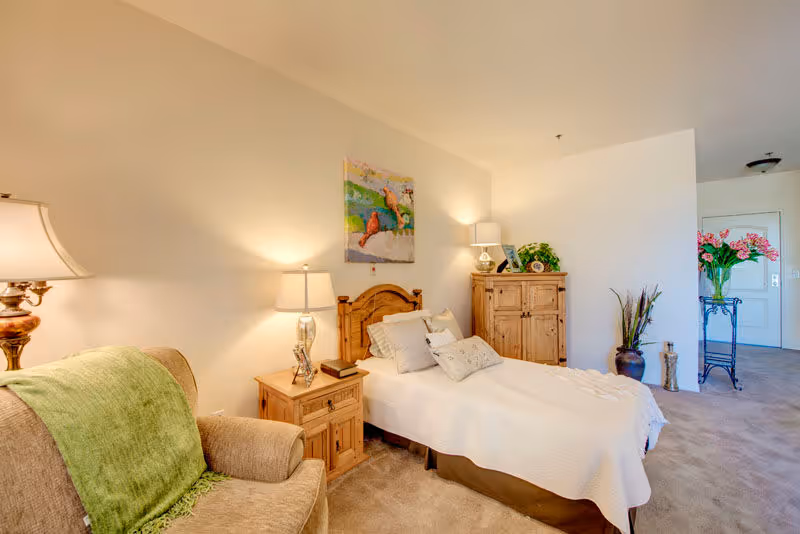 A cozy bedroom with a single bed covered in white bedding and several pillows. Next to the bed is a wooden nightstand with a lamp and a book. There is a wooden dresser against the wall with decorative items and a lamp on top. A comfortable armchair with a green throw blanket is positioned near the bed. The room has beige walls and carpeted flooring, with a colorful painting of birds hanging above the bed. In the background, there is a small table with a vase of pink flowers near the entrance door.