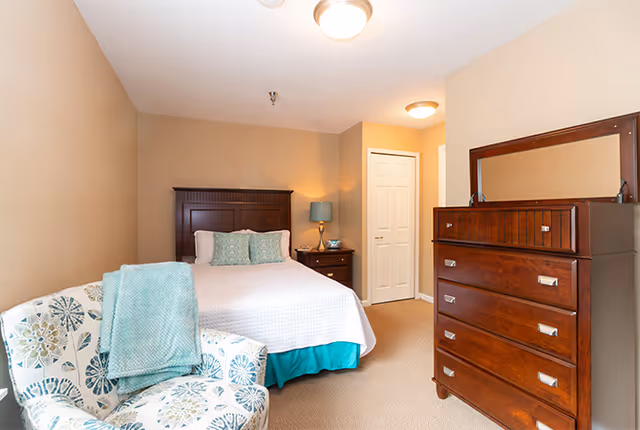 A cozy bedroom with beige walls featuring a bed with a dark wooden headboard, white bedding, and two pillows. Next to the bed is a wooden nightstand with a lamp and a clock. A patterned armchair with a teal throw blanket is positioned in the corner. Across from the bed is a tall wooden dresser with a mirror on top. A closed white door is visible in the background.