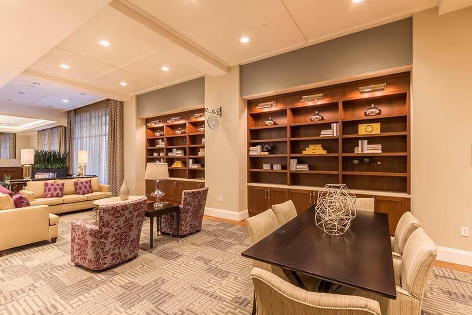 Community lounge with sofas, patterned armchairs, a long table and built-in wooden bookshelves under warm lighting.