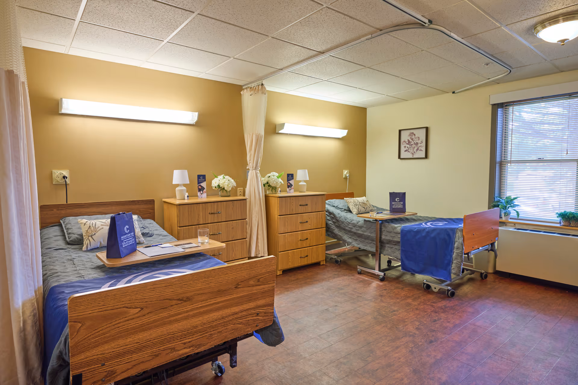A shared patient room in a healthcare facility with two hospital beds separated by a curtain. Each bed has a bedside table with a lamp, flowers, and informational brochures. The room has warm yellow walls, a window with blinds, and wood-patterned flooring.