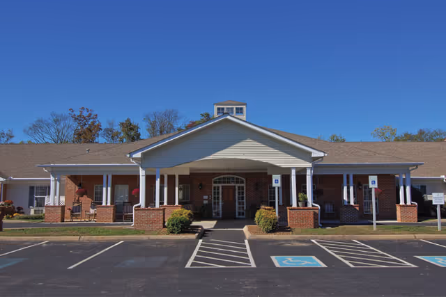 Front exterior view of a single-story brick building with a covered entrance supported by white columns. There are designated handicap parking spaces in front of the building, and the sky is clear and blue.