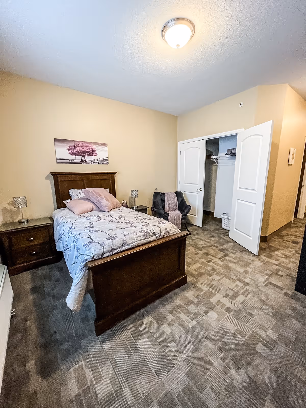 A bedroom with a wooden bed frame and floral bedding, two matching nightstands with lamps, a black chair with a pink throw, an open closet with white double doors, beige walls, and patterned carpet flooring.