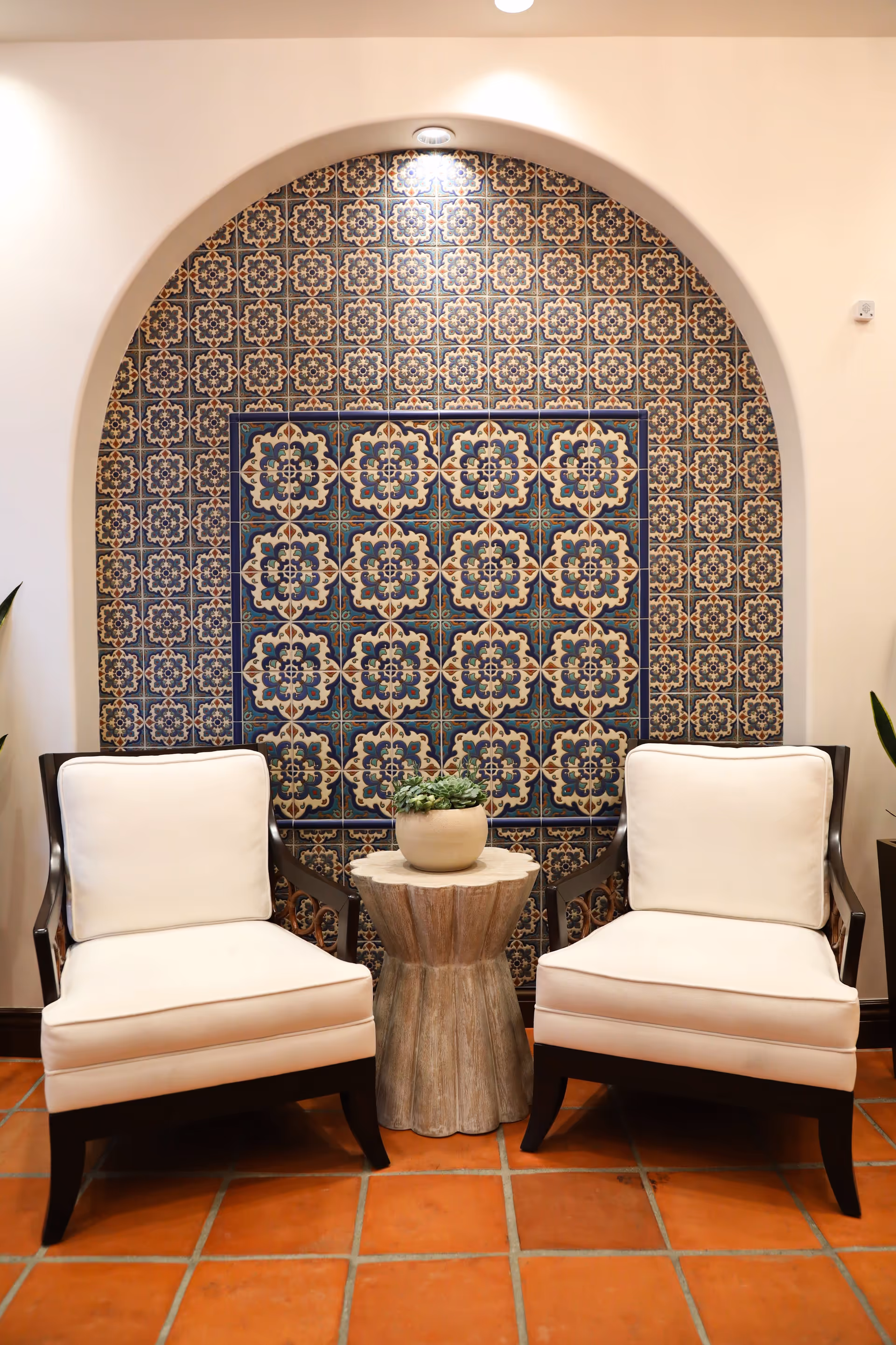 Two white upholstered armchairs flank a small wooden side table with a potted plant in front of an arched decorative tile wall.