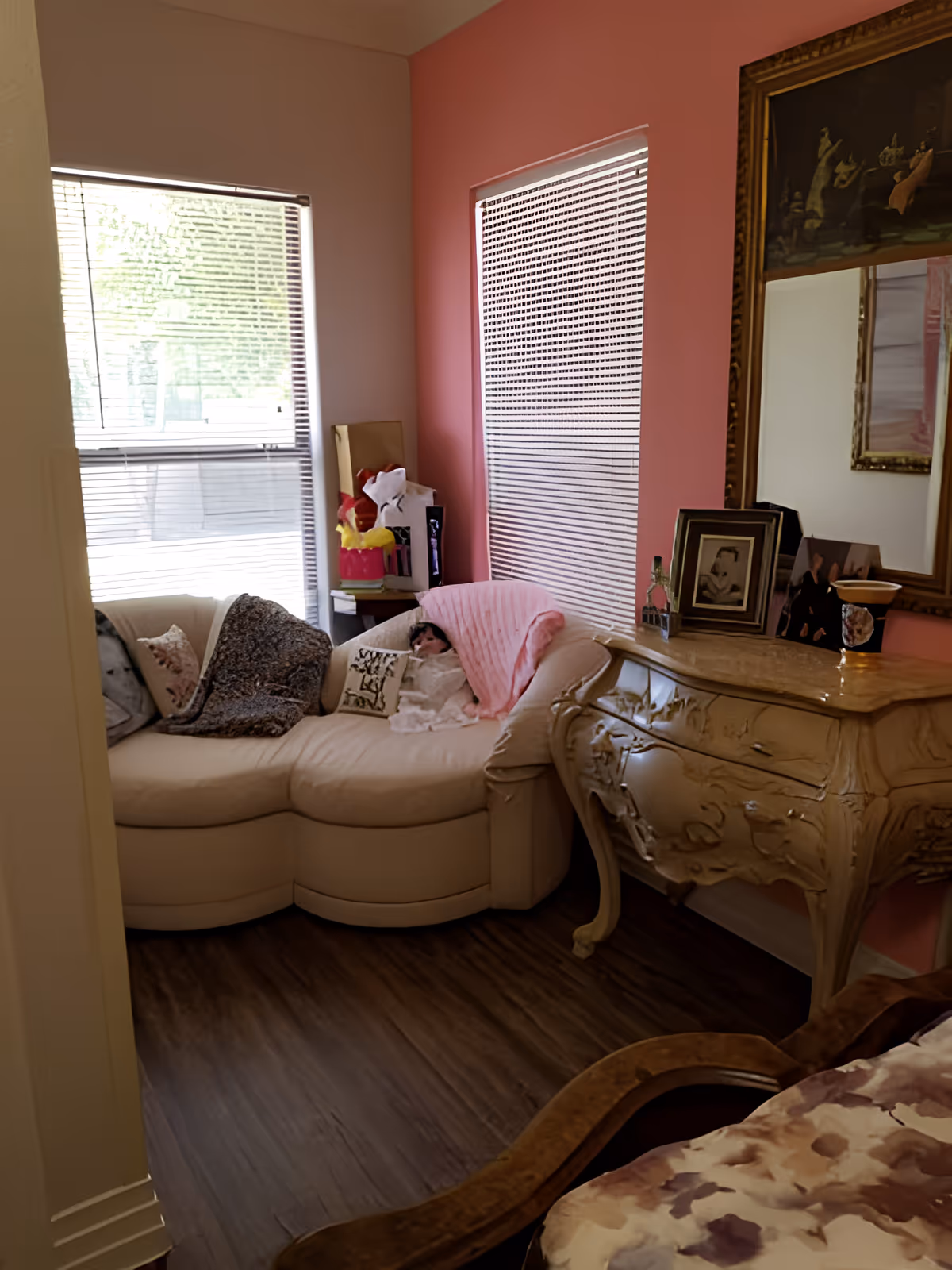 Bedroom corner with a curved cream loveseat, ornate dresser topped with framed photos and a mirror, and two windows with blinds.