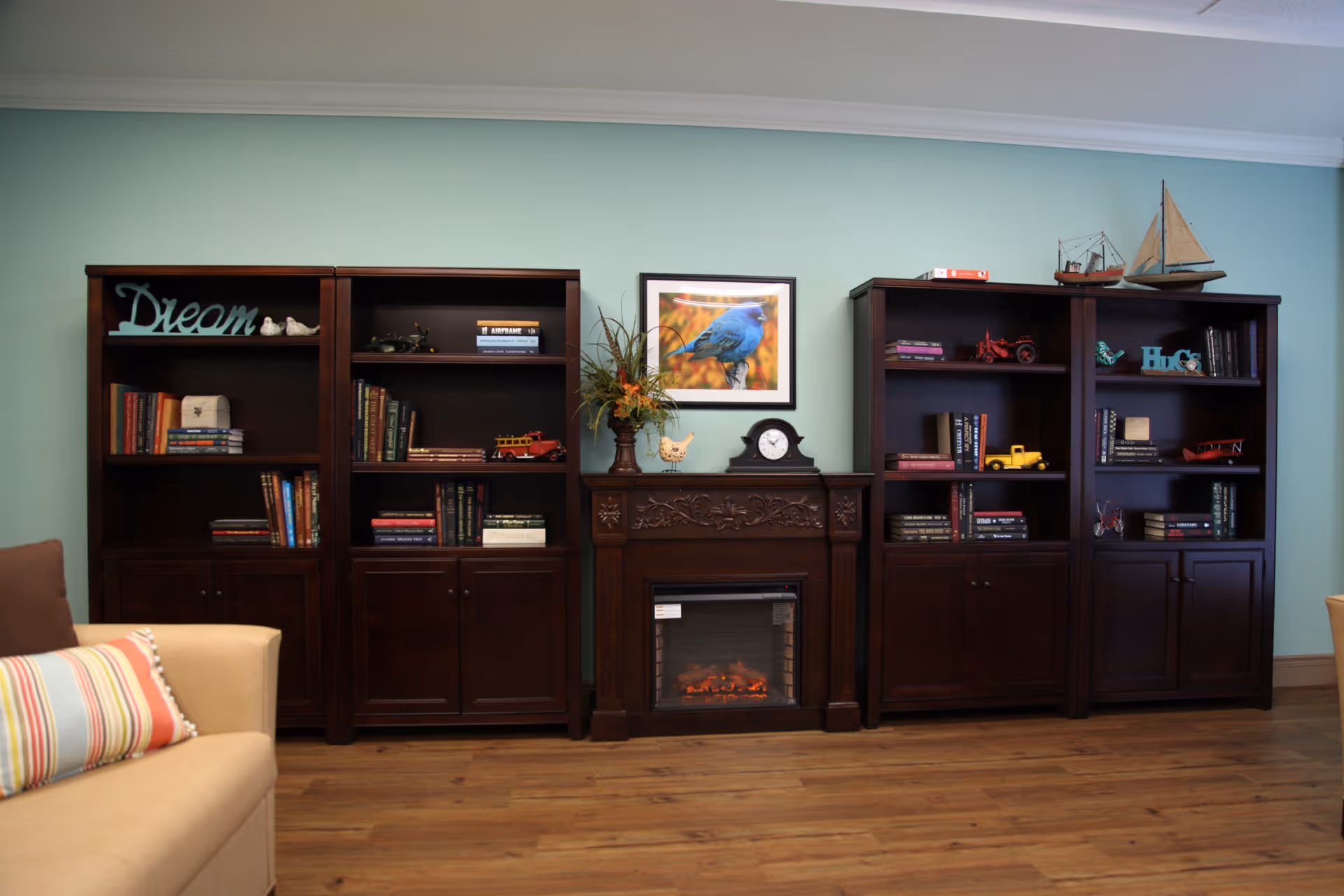 A cozy living room area with three dark wooden bookshelves filled with books and decorative items such as model vehicles, a clock, and small sculptures. In the center is an electric fireplace with a floral arrangement and a framed picture of a blue bird above it. A beige couch with a striped pillow is partially visible on the left side. The walls are painted light green and the floor is wooden.