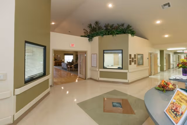 Bright interior reception and hallway area of a senior living facility with a front desk, double doors leading to a seating room, and plants above the partition.