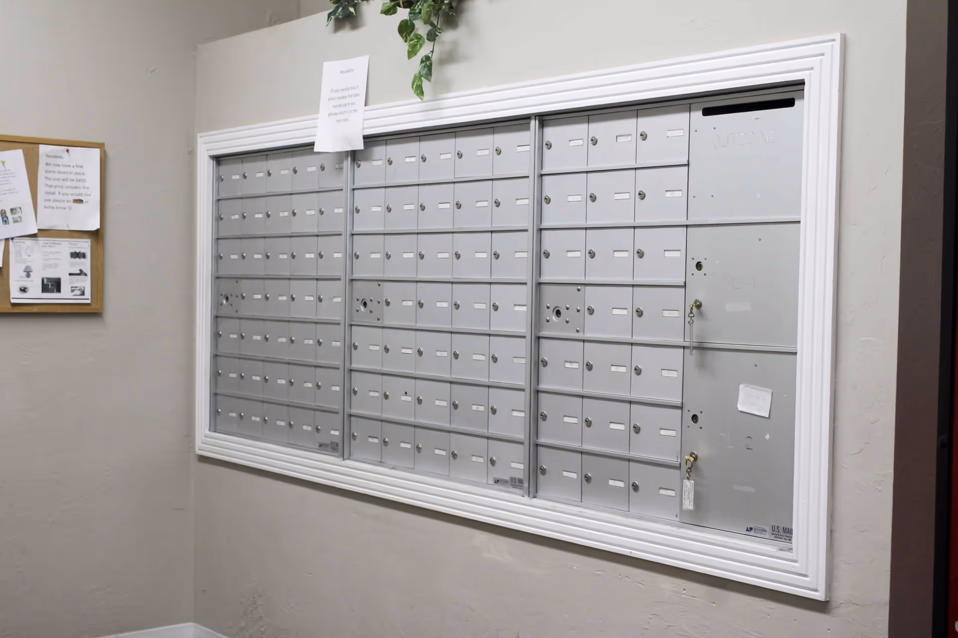 A wall-mounted bank of silver mailboxes with multiple locked compartments, some with keys hanging. Above the mailboxes is a small sign with instructions. To the left, a bulletin board with various notices is visible. The wall and trim are painted in neutral tones.