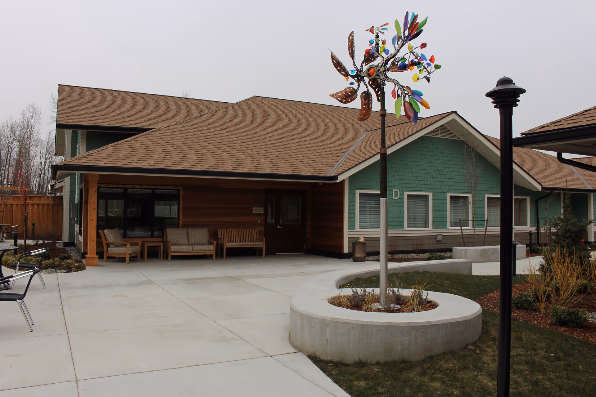 Exterior front courtyard of a single-story senior living building with a covered entrance, seating, a colorful wind sculpture, and paved walkways.